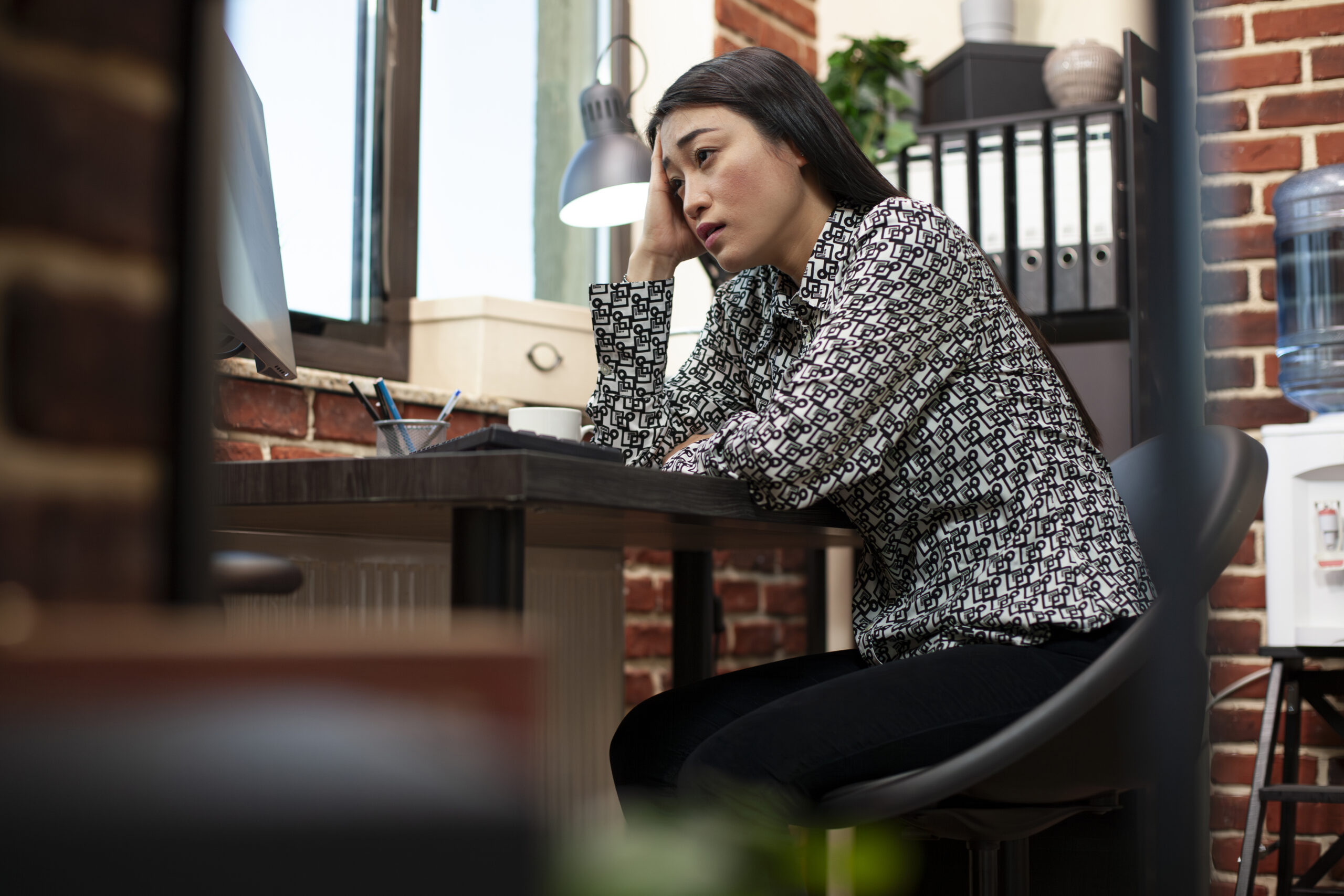 Exhausted female employee seated at desk with computer, visibly tired from financial tasks in startup office. Asian entrepreneur resting on her hand and reviewing company workload in modern workspace.