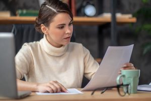 Focused young Caucasian woman sitting at the writing table while scrutinizing documents in her hand