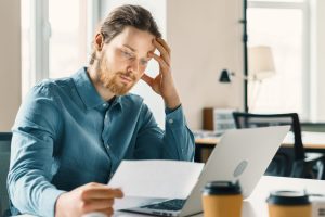 Young upset businessman looking at financial document or bill with frustrated face expression while sitting behind desk with modern laptop computer in office, entrepreneur receiving debt notification